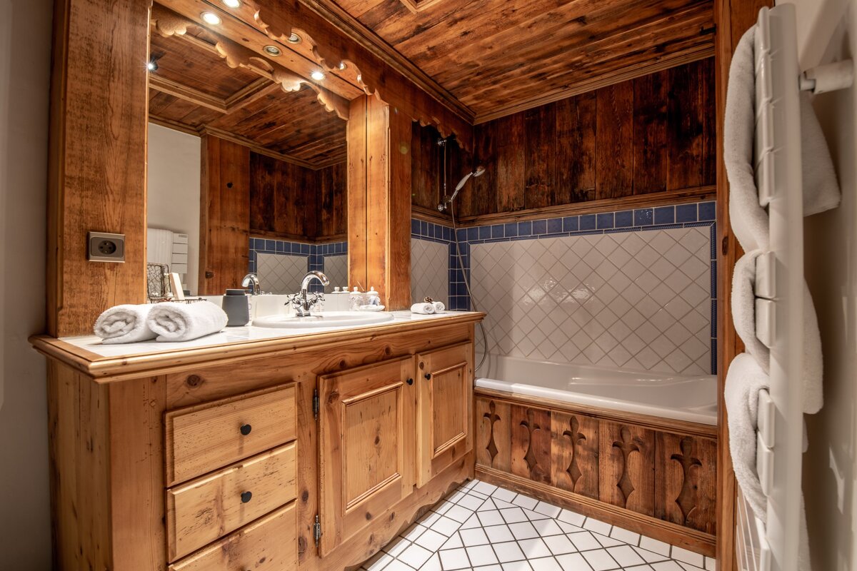 A rustic wooden bathroom with white sink, bathtub, and diamond-patterned white floor/wall tiles bordered by blue, reflecting a warm chalet style.