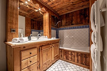 Rustic bathroom with a carved wooden mirror and vanity, a bathtub featuring white diamond tiles with blue trim, and warm wood-paneled walls and ceiling.