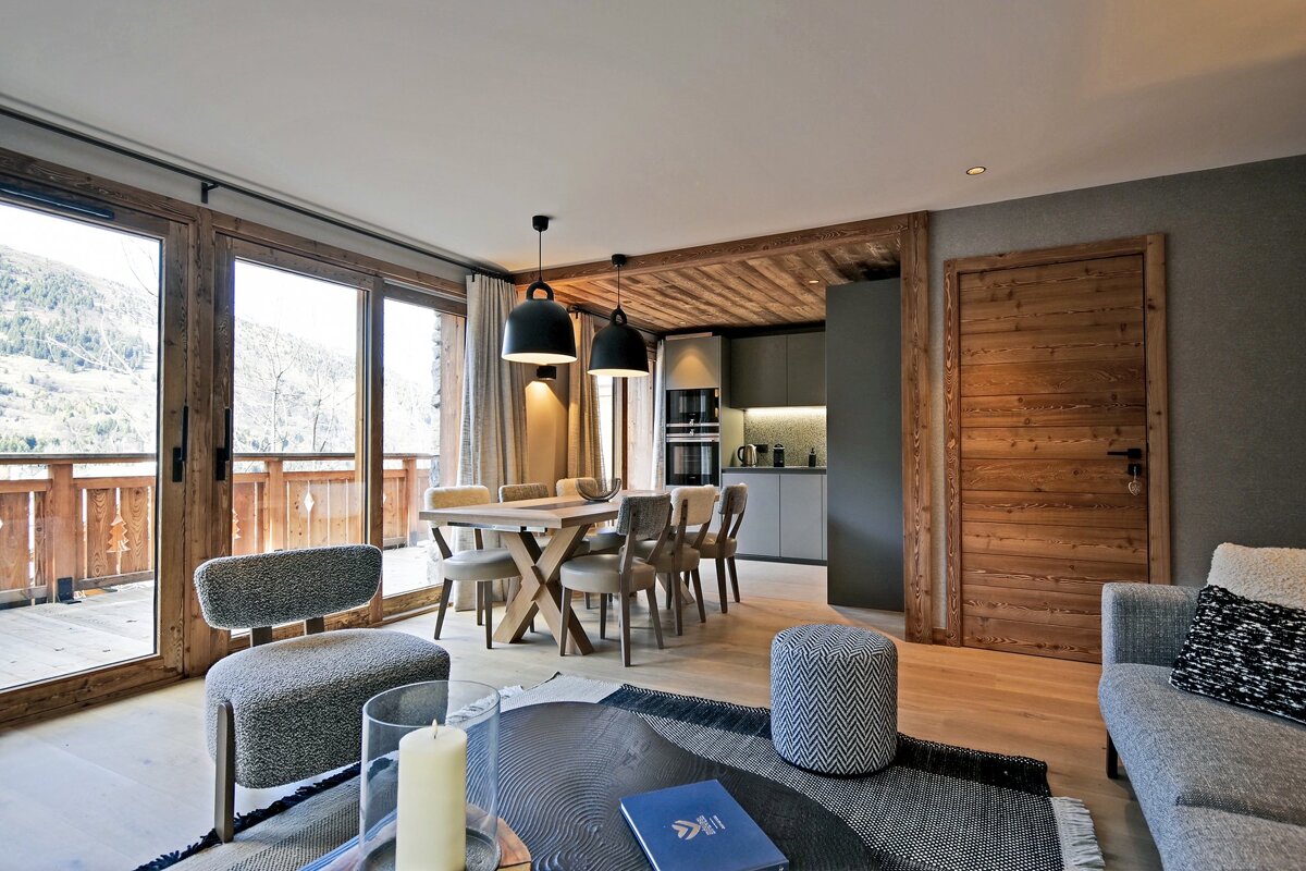 A stylish open-plan living area with a dining table, kitchen, and balcony offering mountain views, featuring modern and chalet-style decor.
