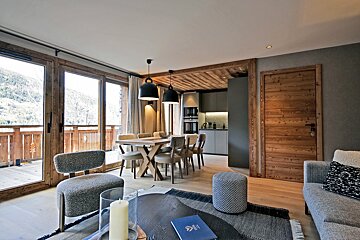 A stylish open-plan living area with a dining table, kitchen, and balcony offering mountain views, featuring modern and chalet-style decor.