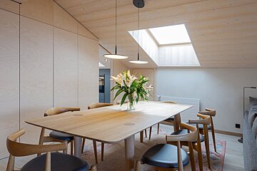 A minimalist wooden dining table with chairs under a skylight, decorated with a vase of lilies.