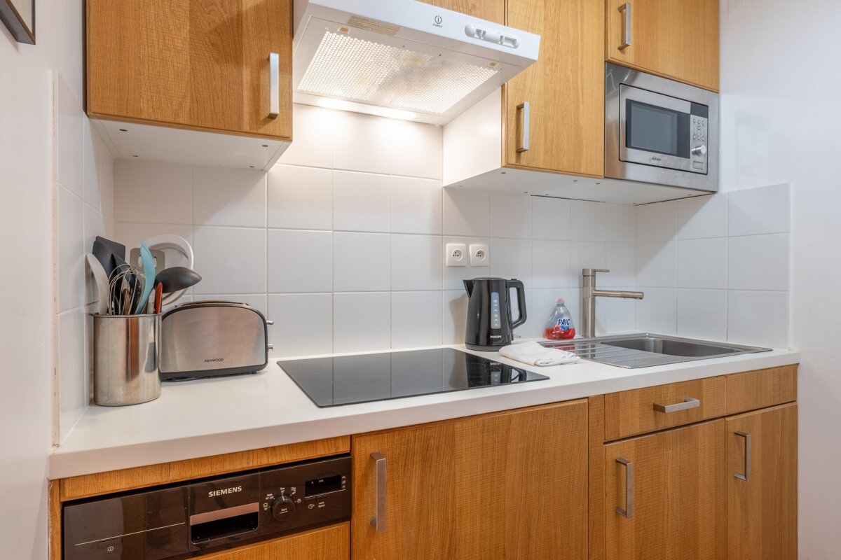 A functional modern kitchen with wooden cabinets, white tiled walls, and appliances like a microwave, hob, toaster, kettle, and sink.