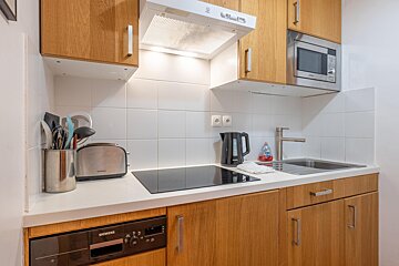 A functional modern kitchen with wooden cabinets, white tiled walls, and appliances like a microwave, hob, toaster, kettle, and sink.