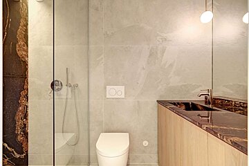 A modern bathroom featuring light grey wall tiles, a glass shower partition, a toilet, and a vanity with a dark marble countertop and sink.
