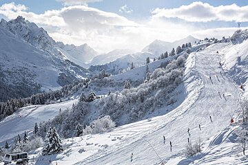 Skiers descend a sunlit slope in a vast, snowy mountain valley with frosted trees and towering peaks under a partly cloudy sky.