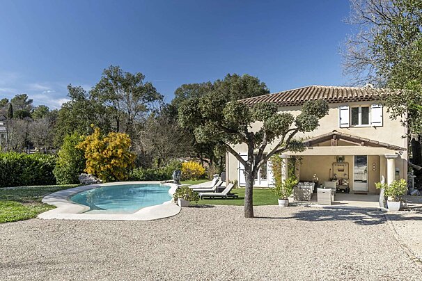 Sunny day view of a light-colored house with a tiled roof, white shutters, and a porch. A freeform pool, gravel area, and lush trees fill the yard.