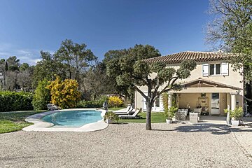 Sunny day view of a light-colored house with a tiled roof, white shutters, and a porch. A freeform pool, gravel area, and lush trees fill the yard.