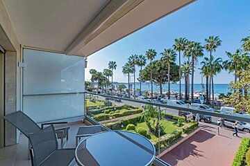 A sunny balcony view with modern furniture overlooking palm trees, a coastal road, and the blue sea.