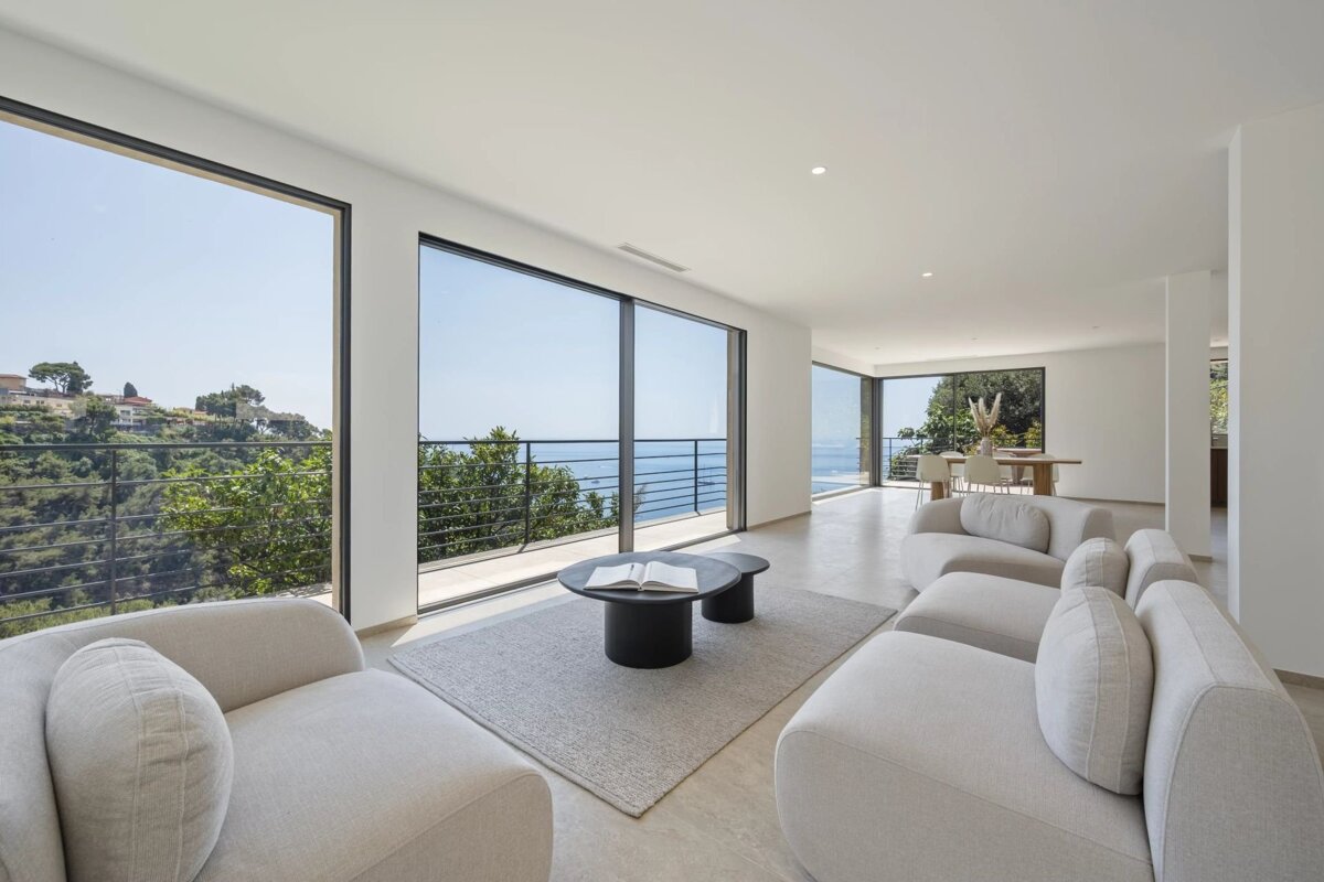 Modern minimalist living room with white sofas, large windows overlooking the ocean, and a neutral color palette.