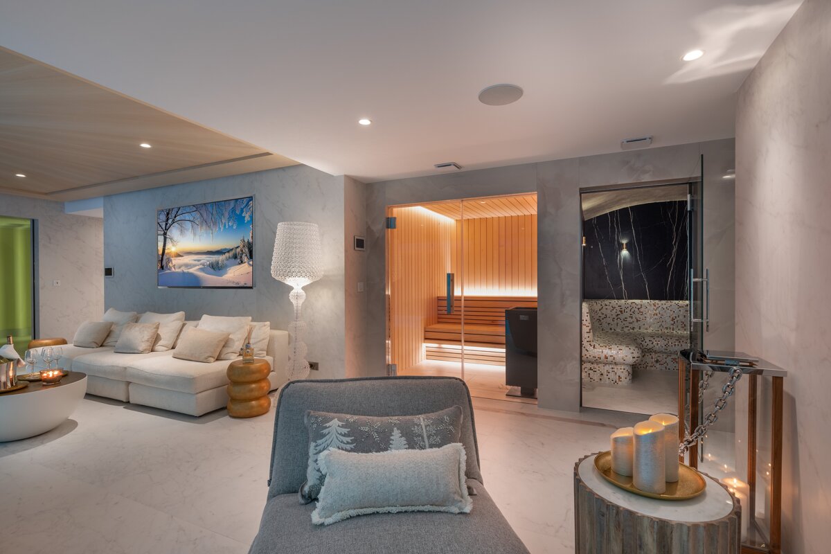 A luxurious spa-like room featuring a wooden sauna, a dark tiled steam room, and a cozy lounge area with a large sofa, chaise, and decorative lighting.