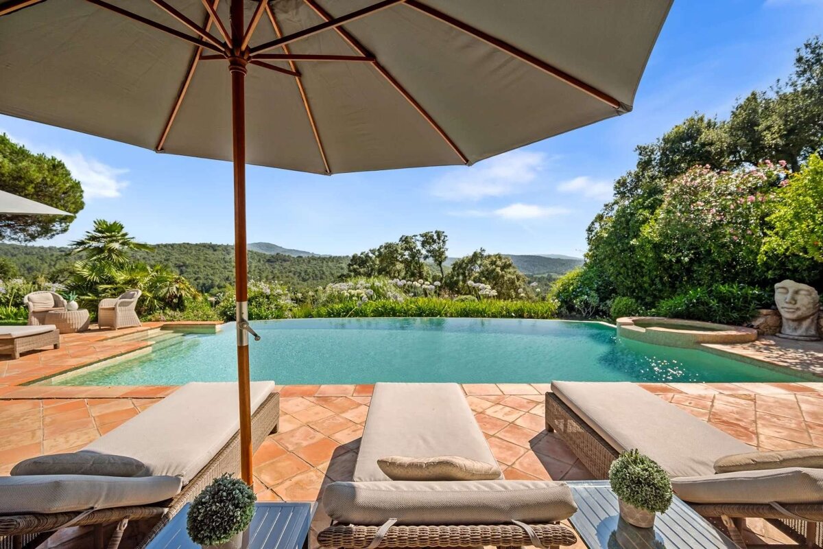 A sunny infinity pool on a terracotta patio, with lounge chairs and an umbrella, offers breathtaking views of rolling green hills and a blue sky.