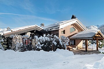 A snow-covered wooden chalet with intricate roof details, surrounded by deep snow and bushes under a clear blue sky, with distant mountains.