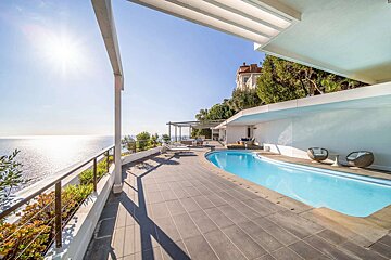 A modern terraced area with a swimming pool, overlooking a sparkling ocean under a bright, sunny sky.