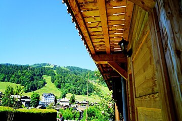 Scenic view of an alpine village and green mountains from the wooden eaves of a rustic chalet under a clear blue sky.