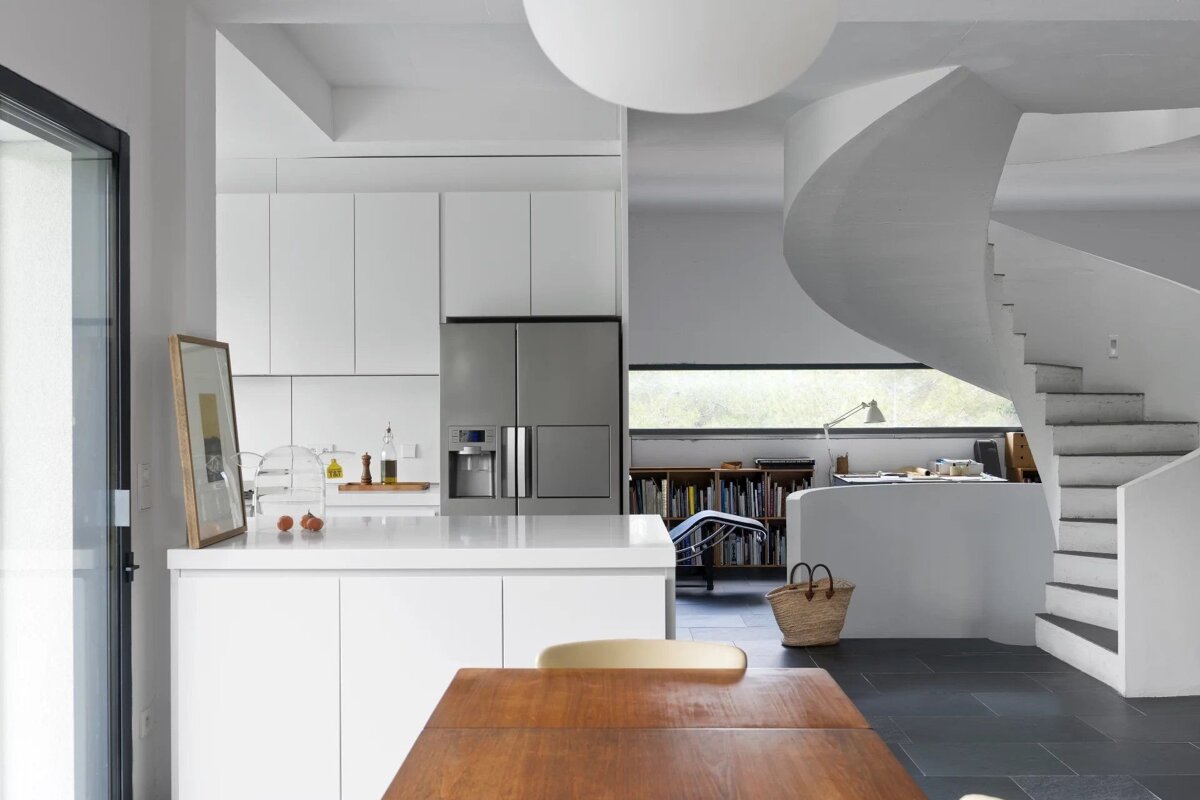 A modern, white interior featuring a minimalist kitchen, a wooden table, and a sweeping spiral staircase leading upstairs.