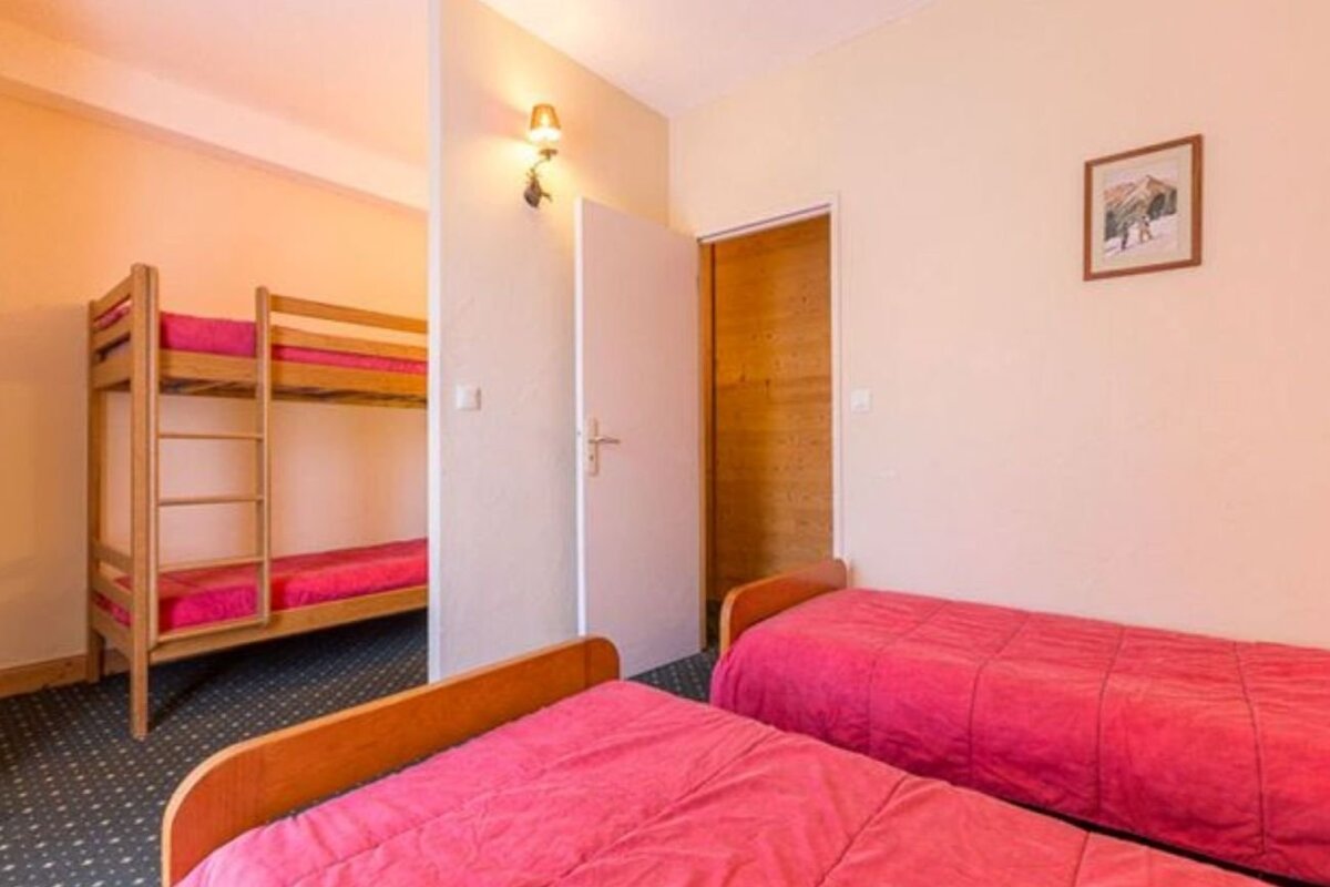 A bedroom with two single beds with pink covers in the foreground and a wooden bunk bed visible behind a partition wall.