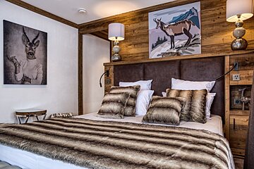 A warm, rustic bedroom features a large bed with faux fur bedding, wooden paneling, a padded headboard, and eclectic animal art.