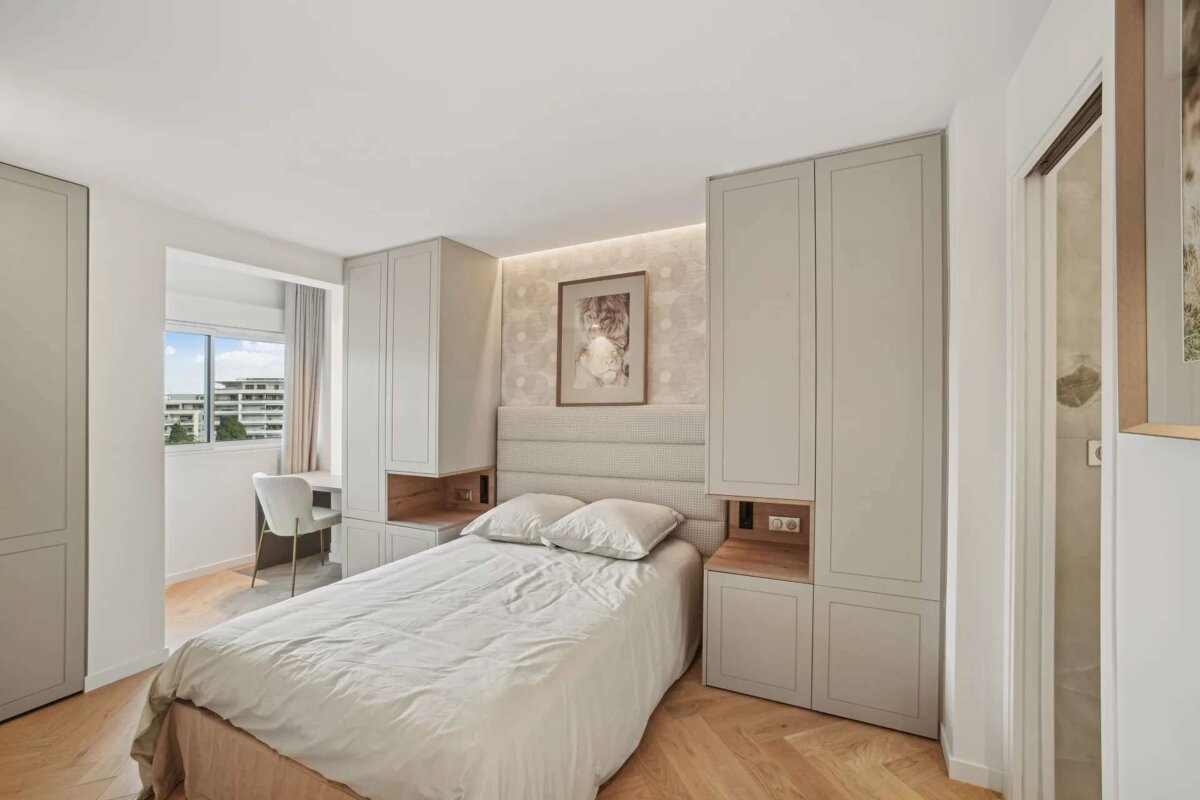 A modern bedroom features a light grey bed flanked by built-in wardrobes, a desk by the window, and light wood herringbone flooring.