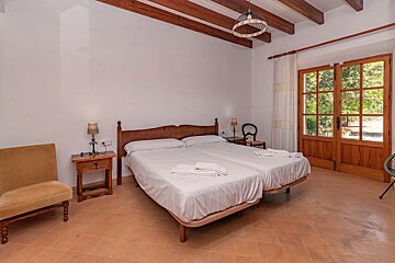 Rustic bedroom with twin beds, exposed wood beams, terracotta floor, and large glass doors leading to greenery.