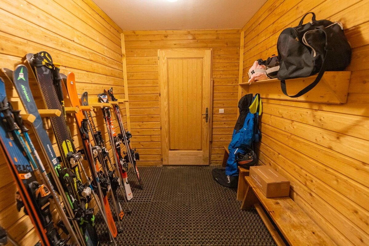 A cozy wooden ski room with numerous skis and poles stored on racks on the left, and ski gear, bags, and clothing on shelves and a bench on the right.