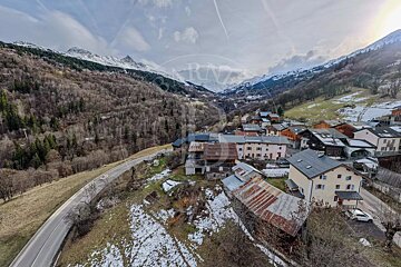 Scenic alpine village in a valley with snow-capped mountains, rustic houses, and a winding road.