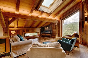 A cozy, rustic wooden chalet living room with exposed beams, skylight, TV, and panoramic mountain views from a large window.
