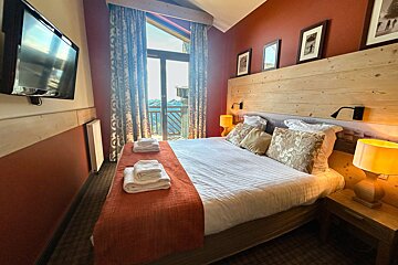 A cozy hotel bedroom with red walls, wood paneling, a large bed with orange throws, and a glass door leading to a balcony.
