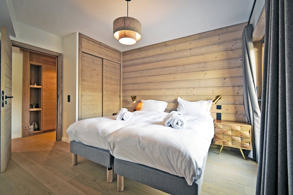 A bright, modern bedroom featuring wood-paneled walls, two beds with white linens, light wooden floors, and simple, cozy decor.