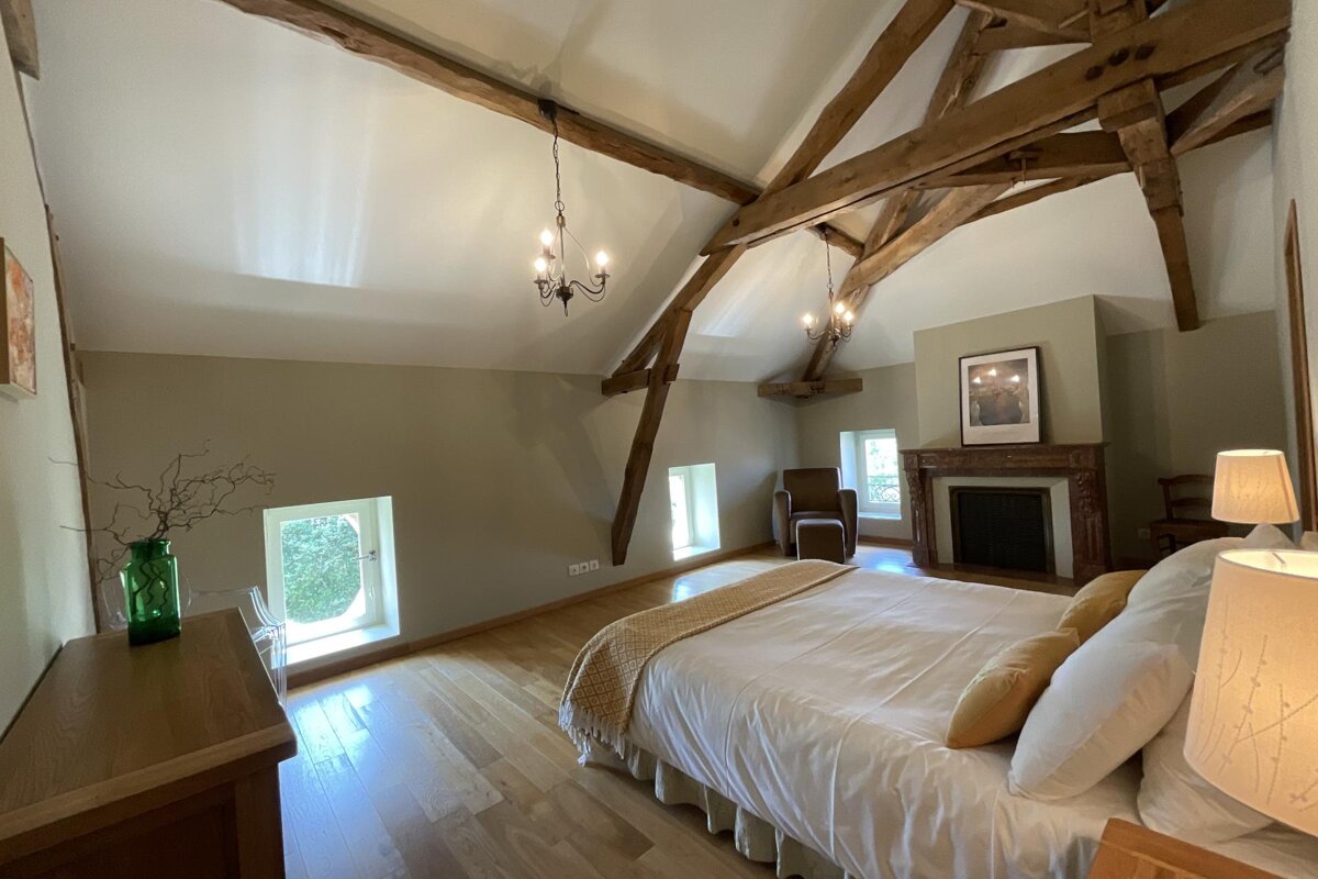 A rustic chic bedroom featuring exposed wooden beams, a high vaulted ceiling, a stately fireplace, hardwood floors, and a cozy bed with white linens.