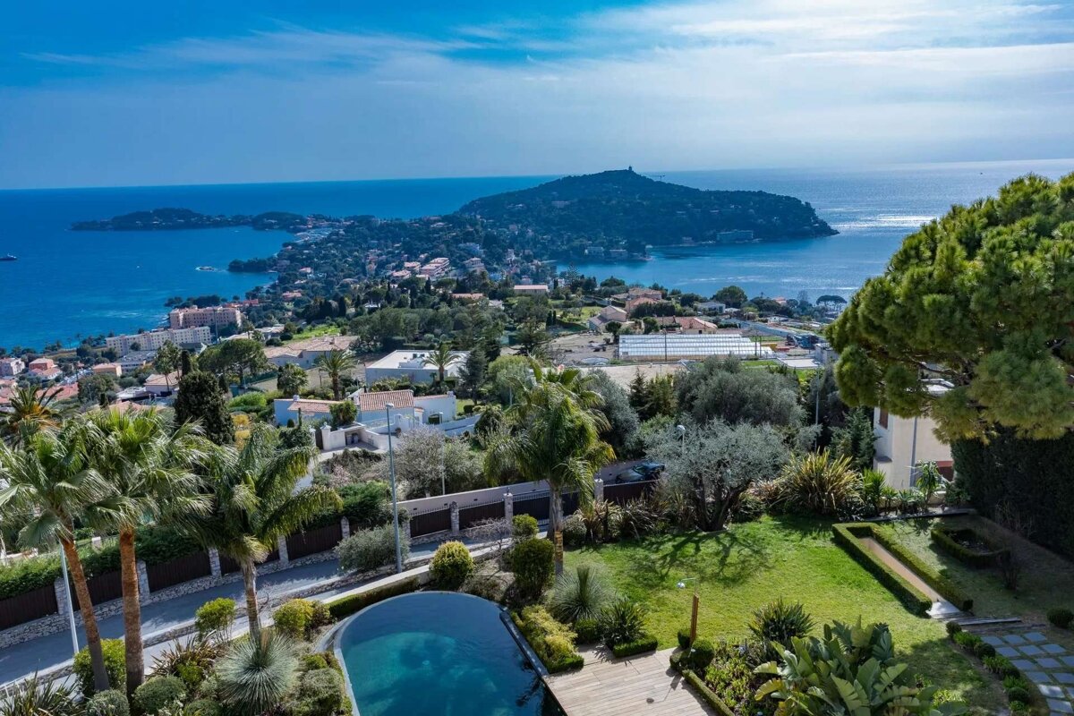 Elevated view of a beautiful Mediterranean coastal town, featuring a peninsula, blue sea, lush gardens, and a private swimming pool under a bright sky.