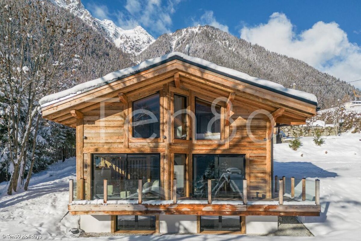 A modern wooden chalet with a snowy roof, surrounded by snow, against a stunning backdrop of snow-covered mountains under a bright blue sky.