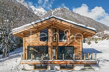 A modern wooden chalet with a snowy roof, surrounded by snow, against a stunning backdrop of snow-covered mountains under a bright blue sky.