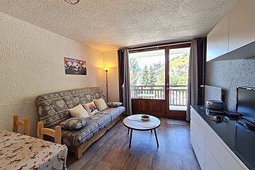A cozy, brightly lit living room with a patterned sofa, small coffee table, and a balcony revealing a beautiful mountain and tree view.