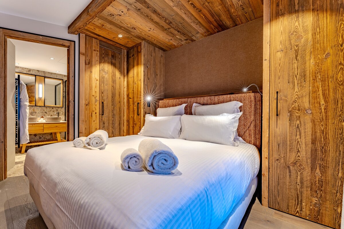 Cozy wooden chalet bedroom with a neatly made bed, rolled towels, and a glimpse into the en-suite bathroom.