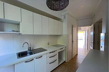 A bright, modern white kitchen with sleek cabinets, a sink, stovetop, and a wicker light fixture. A hallway is visible through an open doorway.