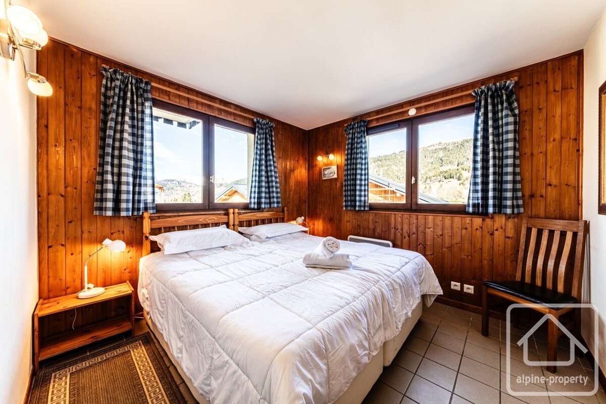 Cozy, wood-paneled bedroom with a white bed and plaid curtains. Windows frame a scenic mountain view and sunny alpine landscape.