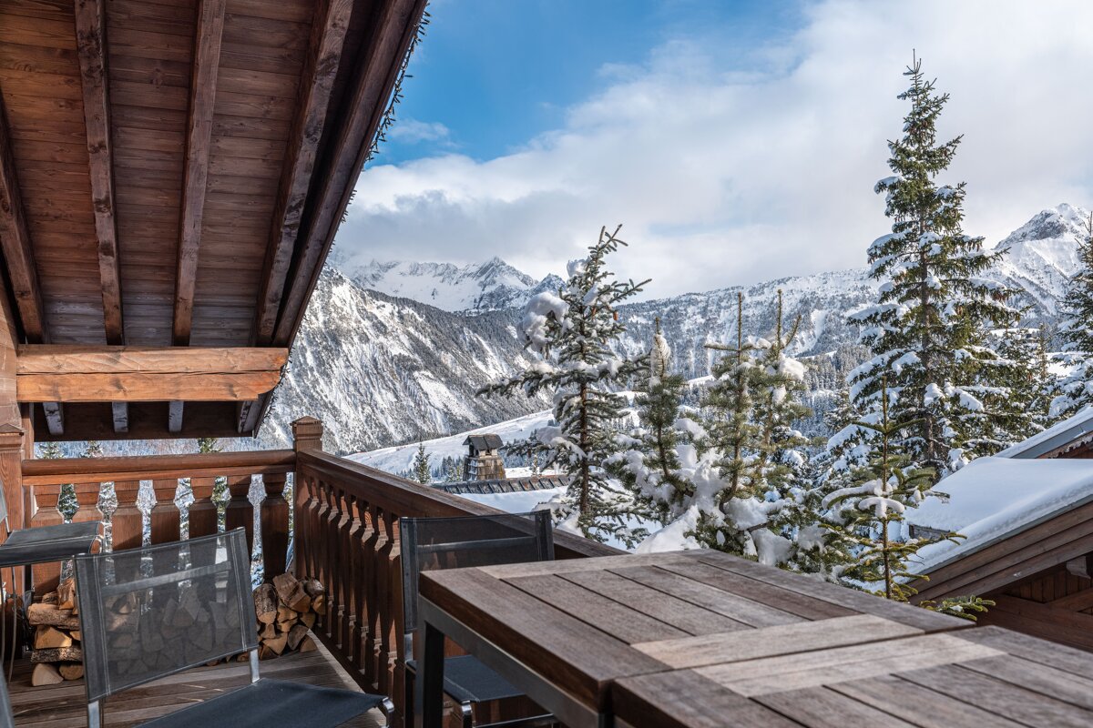 A wooden chalet balcony offers scenic views of snow-covered pine trees and majestic mountains.