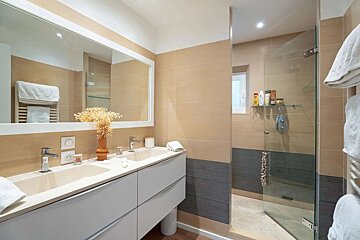 A modern bathroom featuring a double vanity with a large mirror and a glass-enclosed shower. Neutral beige and gray tiles create a clean, minimalist aesthetic.