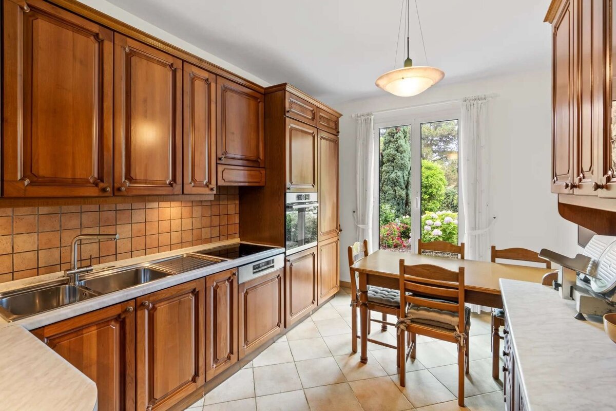 A cozy kitchen with traditional wooden cabinets, a dining table, tiled floor, and a bright garden view through large windows.