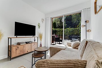 A bright, modern living room with a beige sofa, wooden TV stand, and sliding glass doors opening to a green garden patio.