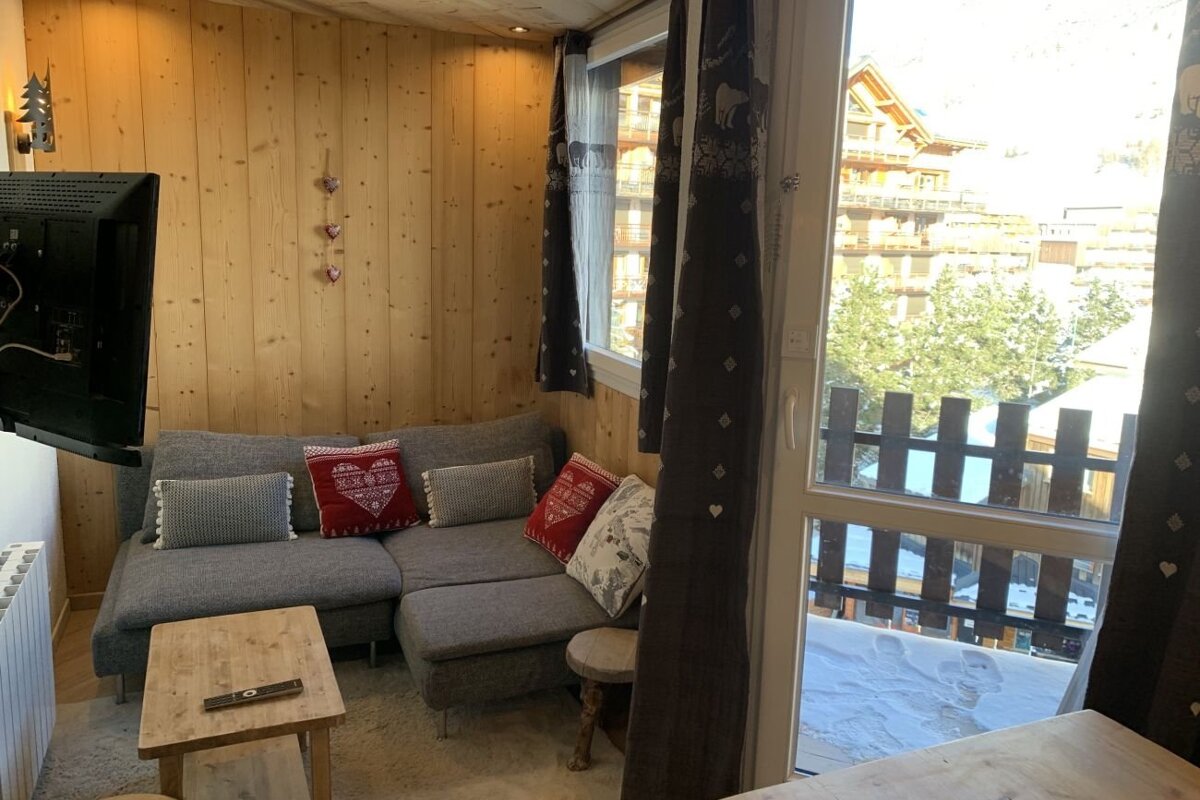 Cozy chalet living room with wood-paneled walls, a grey sofa, and a glass door opening to a snowy mountain resort.