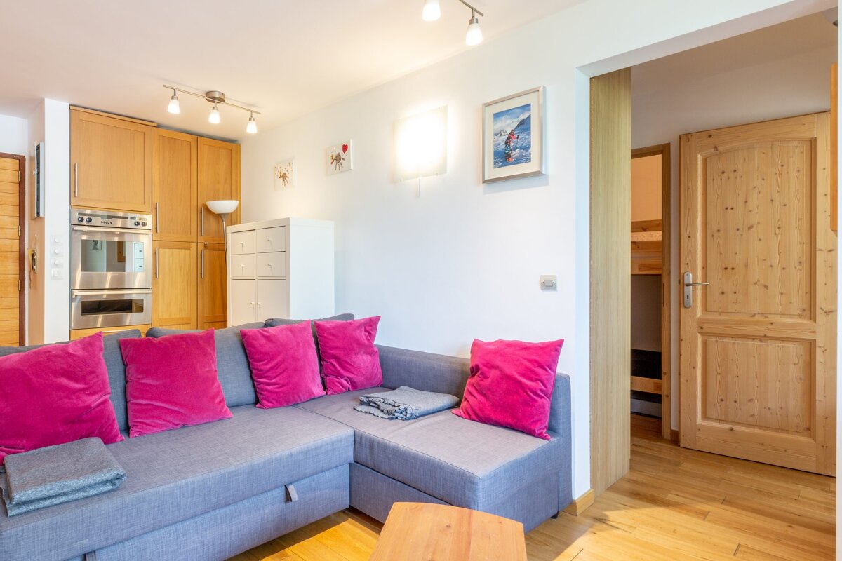 Cozy living room with a grey sectional sofa decorated with bright pink pillows, wooden floors, and kitchen cabinets in the background.