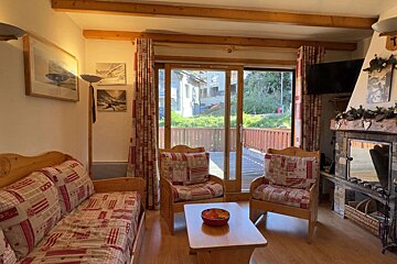 Cozy living room with wooden furniture, a stone fireplace, and large glass doors opening to a balcony. Patchwork textiles and mountain art complete the chalet feel.