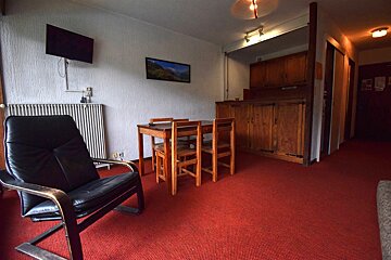 Interior of a room with red carpet, a black leather armchair, a wooden dining set, and a kitchen area in the background.
