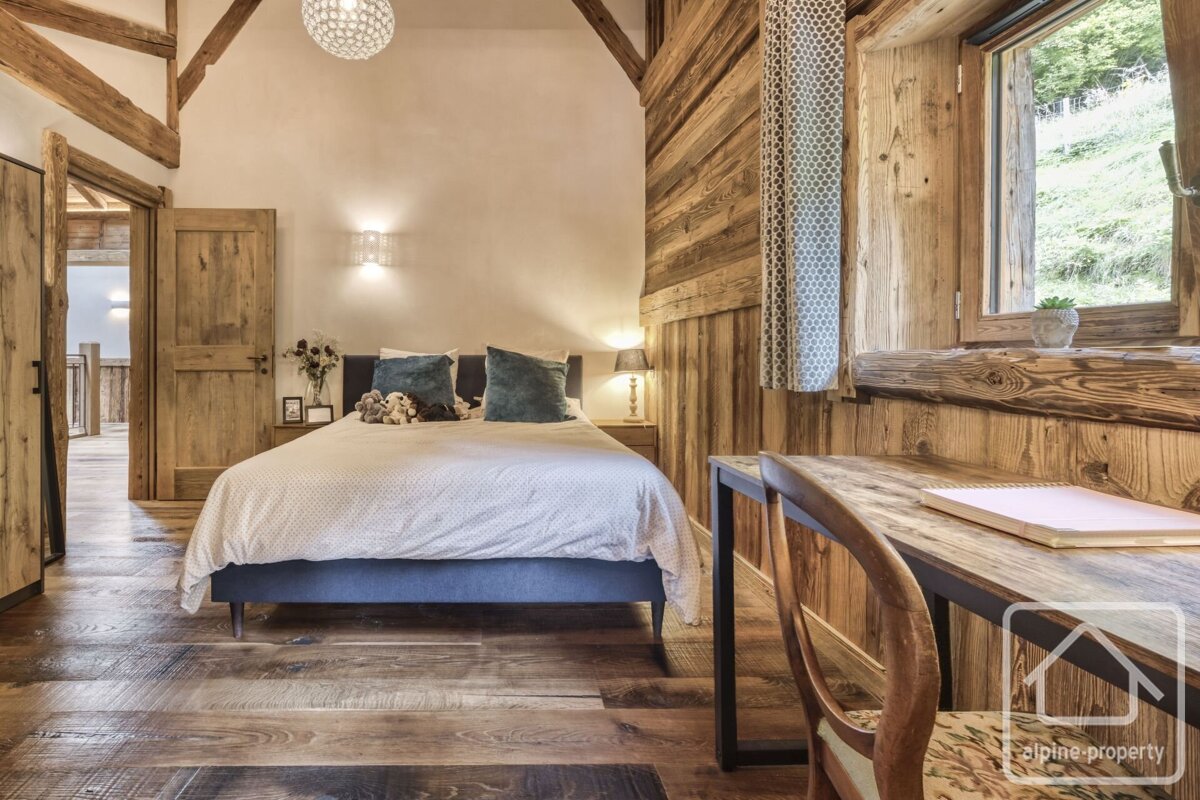 A cozy, rustic bedroom with extensive wood paneling, floor, and beams. A comfy bed and a desk by a window with a green view complete the inviting space.