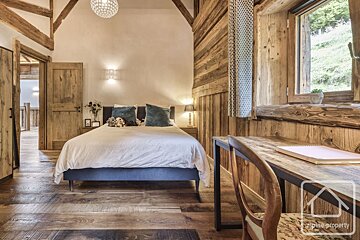 Rustic chalet bedroom with wood paneling, exposed beams, a cozy bed with blue cushions, and a desk by a window overlooking nature.