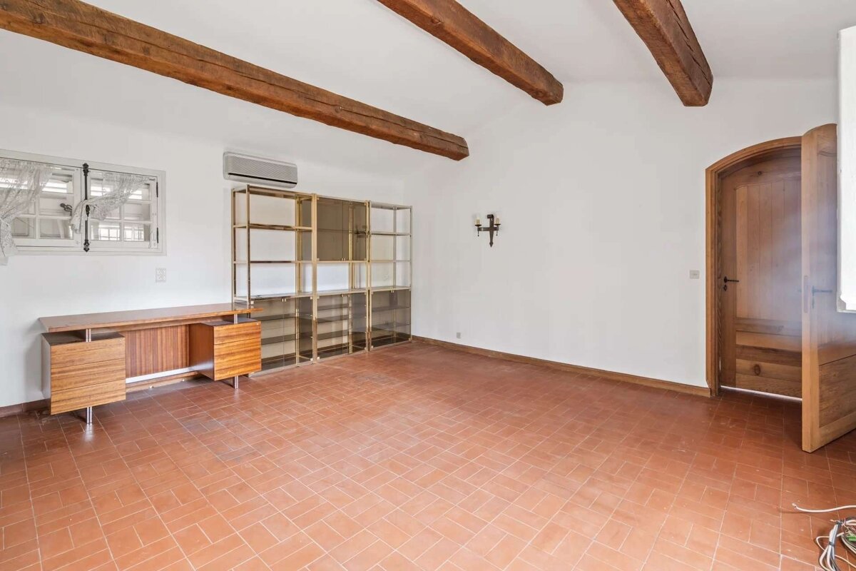 A room with white walls, exposed wooden ceiling beams, and terracotta floor. It features a modern desk, large glass shelving, a window, and an arched wooden door.