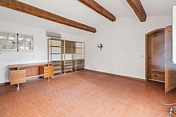 A room with white walls, exposed wooden ceiling beams, and terracotta floor. It features a modern desk, large glass shelving, a window, and an arched wooden door.