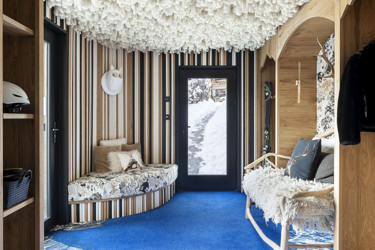 A chic ski chalet entrance with vibrant blue carpet, striped walls, and a fluffy white ceiling. Benches, ski gear, and a snowy exterior create a cozy alpine welcome.