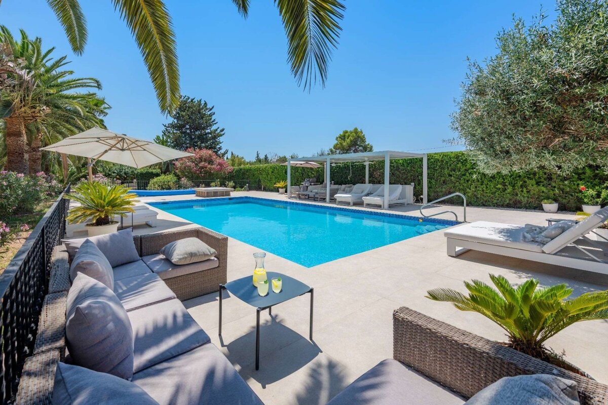 A bright, sunny pool area with a sparkling blue pool, comfortable outdoor lounge furniture, sun loungers, and tropical greenery under a clear sky.